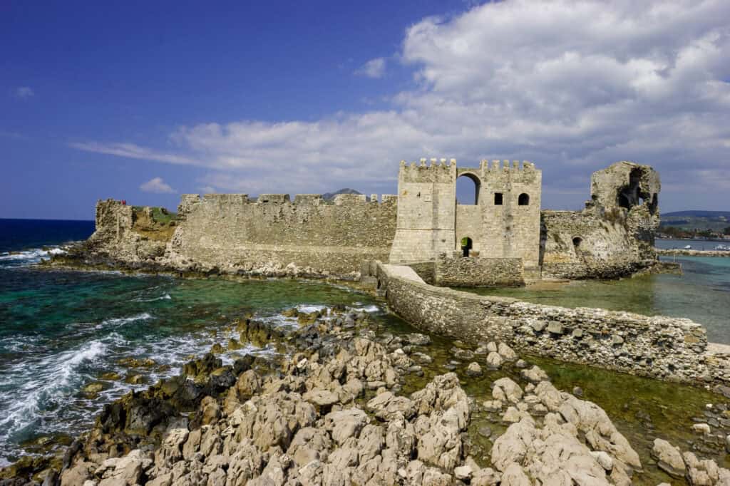 A stone fortress juts out into the turquoise sea under a bright blue sky with fluffy white clouds. The ancient structure, with its crenellated walls and arched openings, is partially surrounded by water and rocky shores.