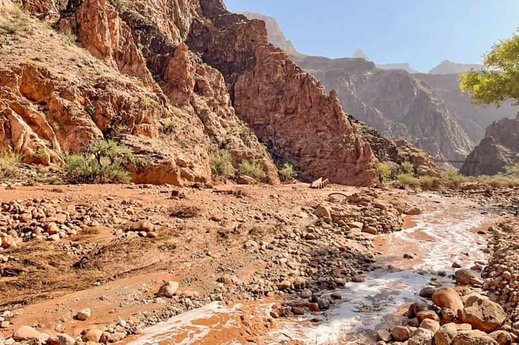 a muddy and rocky creekbed with a canyon and bridge over the river in the background